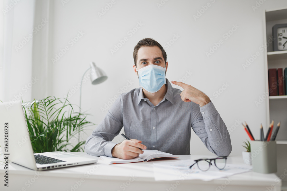 Excited young business man in gray shirt sit at desk work on laptop in light office on white wall background. Achievement business career concept. Writing notes in notebook point on sterile face mask. Excited young business man in gray shirt sit at desk work on laptop in light office on white wall background. Achievement business career concept. Writing notes in notebook point on sterile face mask.