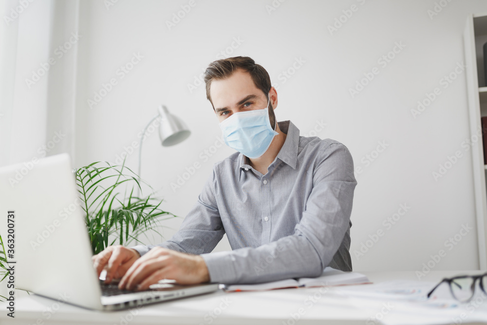Successful young bearded business man in gray shirt sterile face mask sitting at desk in light office on white wall background. Achievement business career concept. Working on laptop pc computer. Successful young bearded business man in gray shirt sterile face mask sitting at desk in light office on white wall background. Achievement business career concept. Working on laptop pc computer.