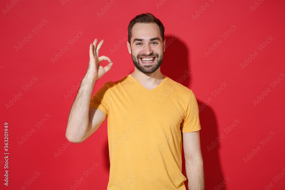 Smiling funny young bearded man guy in casual yellow t-shirt posing isolated on red wall background studio portrait. People sincere emotions lifestyle concept. Mock up copy space. Showing OK gesture. Smiling funny young bearded man guy in casual yellow t-shirt posing isolated on red wall background studio portrait. People sincere emotions lifestyle concept. Mock up copy space. Showing OK gesture.