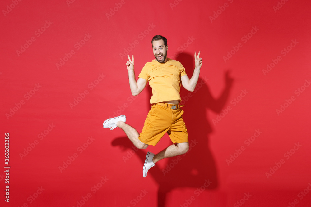Excited young bearded man guy in casual yellow t-shirt posing isolated on red background studio portrait. People sincere emotions lifestyle concept. Mock up copy space. Jumping showing victory sign.