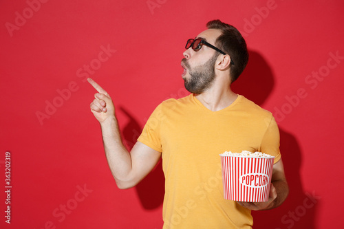 Shocked young man guy in casual yellow t-shirt 3d glasses isolated on red background. People in cinema, lifestyle concept. Watching movie film holding bucket of popcorn pointing index finger aside up.
