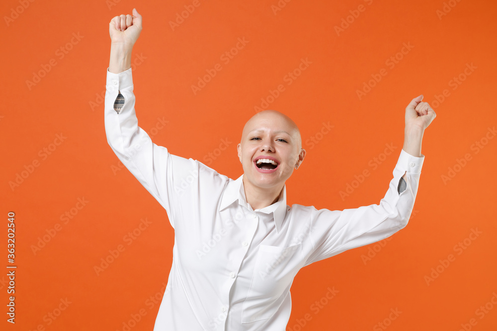 Fototapeta premium Joyful young bald business woman girl in white shirt posing isolated on orange background studio portrait. Achievement career wealth business concept. Mock up copy space. Clenching fists like winner.