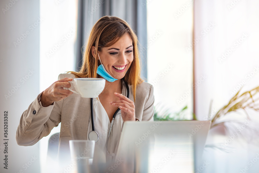 Online doctor with a protective mask tutoring over the internet. Drinking morning coffee. Businesswoman wearing face mask while working on a computer and drinking coffee in the office. Online doctor with a protective mask tutoring over the internet. Drinking morning coffee. Businesswoman wearing face mask while working on a computer and drinking coffee in the office.