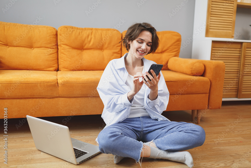 Smiling young woman girl in casual clothes sitting on floor near couch spending time in living room at home. Rest relax good mood leisure lifestyle concept. Using laptop pc computer, mobile phone. Smiling young woman girl in casual clothes sitting on floor near couch spending time in living room at home. Rest relax good mood leisure lifestyle concept. Using laptop pc computer, mobile phone.