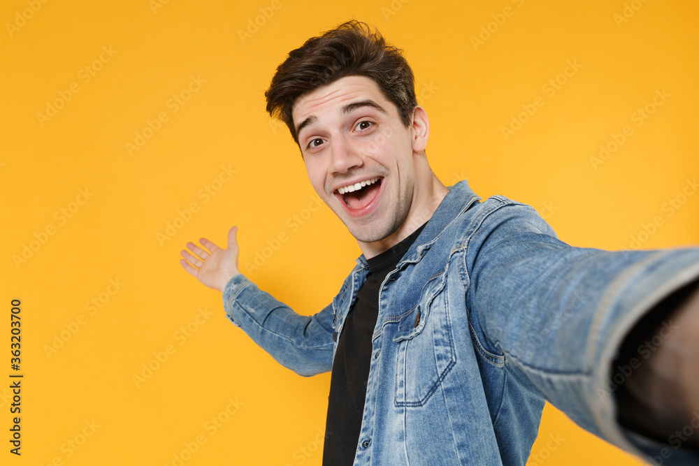 Close up of excited young man guy 20s in casual denim jacket posing isolated on yellow background. People lifestyle concept. Mock up copy space. Doing selfie shot on mobile phone, pointing hand aside. Close up of excited young man guy 20s in casual denim jacket posing isolated on yellow background. People lifestyle concept. Mock up copy space. Doing selfie shot on mobile phone, pointing hand aside.