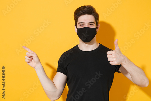 Young guy in casual black t-shirt face mask isolated on yellow background studio. Epidemic pandemic coronavirus 2019-ncov sars covid-19 flu virus concept. Showing thumb up pointing index finger aside.