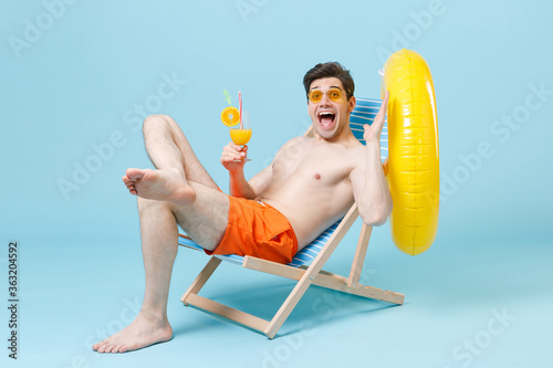 Surprised young man in orange shorts glasses sit on deck chair isolated on blue background. People summer vacation rest lifestyle concept. Mock up copy space. Hold glass of cocktail, spreading hands.