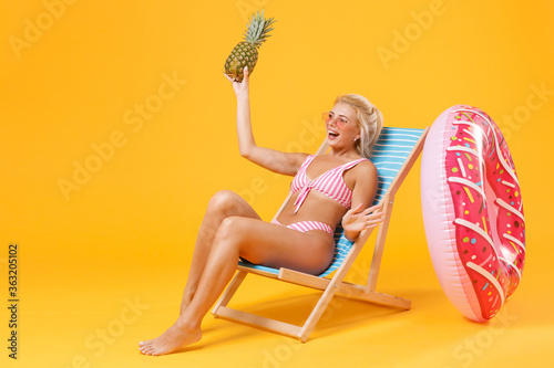 Cheerful young woman girl in pink striped swimsuit glasses sit on deck chair isolated on yellow background. People summer vacation rest lifestyle concept. Mock up copy space. Hold pineapple fruit.