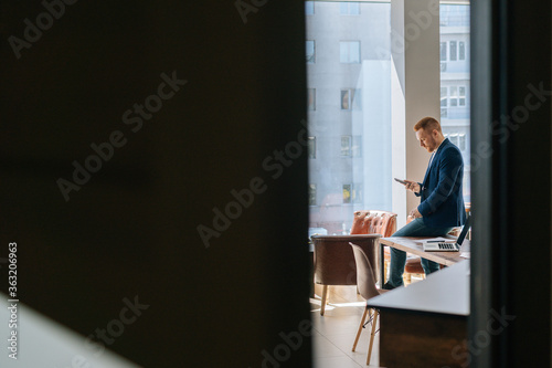Remote view of skilled young man wearing fashion casual clothing is using mobile phone in modern office room near wooden desk on background of large window. Concept of office working.
