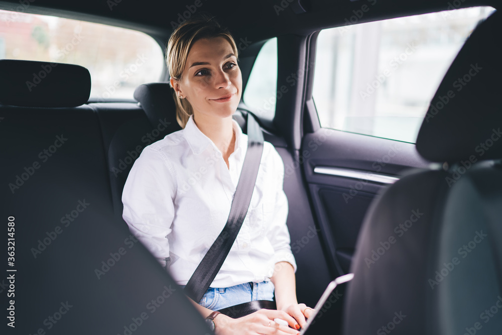 Prosperous female blogger smiling while using car service transportation for getting to own destination in city, happy Caucasian hipster girl with security belt sitting at backseat and smiling