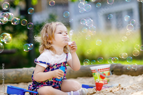 Beautiful little blonde toddler girl having fun with blowing soap bubble blower. Cute adorable baby child playing on playground on sunny summer day. Happy active funny healthy kid