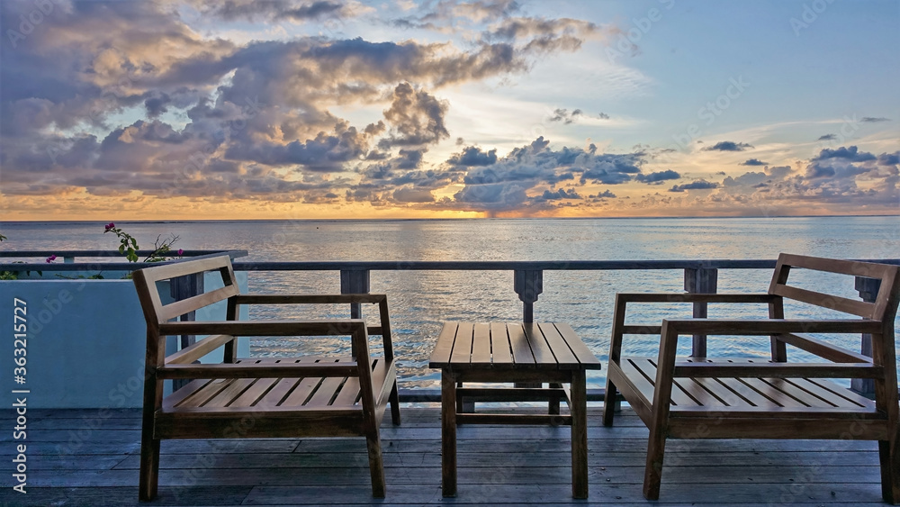 On a wooden platform above the ocean are a table and  chairs. The sun has already sunk over the horizon. The sky and picturesque clouds are painted in gold, orange, lilac, blue tones. Maldives.

