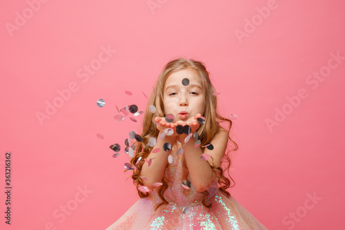 a little girl in an elegant dress blows confetti on a pink background, holiday concept