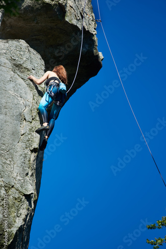 Vertical snapshot of female rock-climber, climbing up a cliff, wearing blue leggings and a black top in safety harness on a sunny day, clear blue sky on background. Low angle view