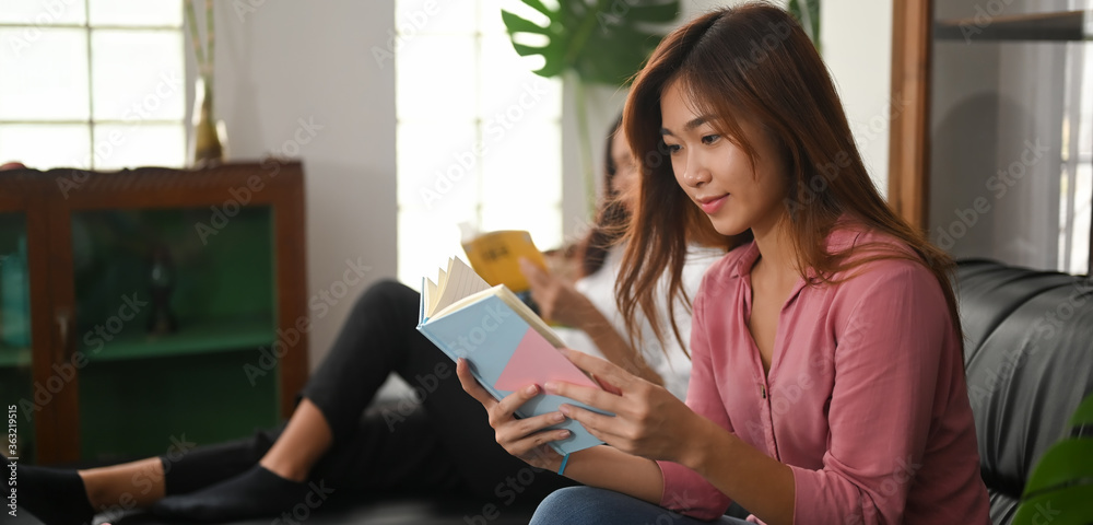 Young Asian women are reading a book while sitting together on a black leather sofa. Young Asian women are reading a book while sitting together on a black leather sofa.