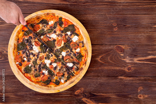 Top view of male hand which cutting the pizza with a kitchen knife. Delicious pizza with cherry, spinach, mozzarella, feta, olive and mushrooms on a wood platter which is on wooden rustic table