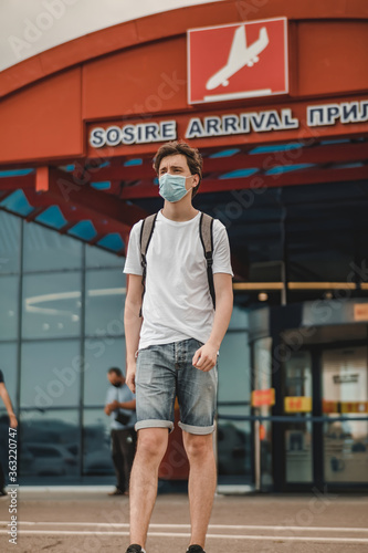 Man dressed in white t-shirt, jeans shorts, black shoes, backpack and surgical mask walking away from airport