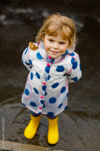 Fototapeta Naklejka Na Ścianę i Meble -  Little toddler girl wearing yellow rain boots, running and walking during sleet on rainy cloudy day. Cute happy child in colorful clothes jumping into puddle, splashing with water, outdoor activity