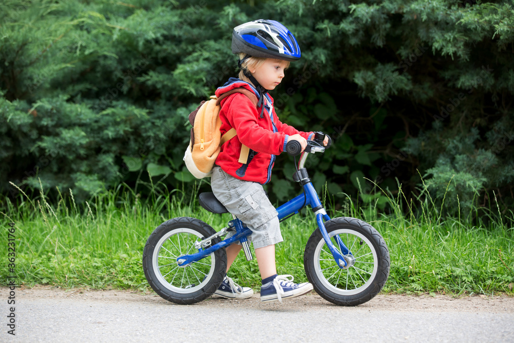 Naklejka premium Cute toddler boy with blue helmet, riding balance bike on the street