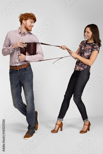 Man and woman model in the studio advertise leather bag. They are dressed in casual clothes jeans, shirts, shoes. Isolated background. A woman is pulling his bag. Smiling. Fool around