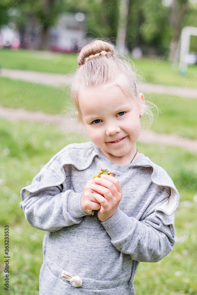 Little blond girl with blue eyes, hair arranged in a bun, wearing a gray dress, close-up portrait, looking into the camera, smiling, bored, playing on a lawn, sitting on the machine tires. Preschool Little blond girl with blue eyes, hair arranged in a bun, wearing a gray dress, close-up portrait, looking into the camera, smiling, bored, playing on a lawn, sitting on the machine tires. Preschool