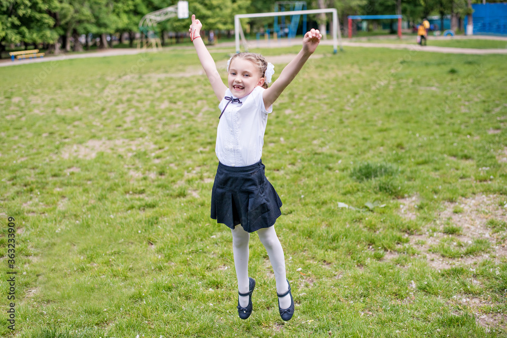 Little blonde schoolgirl in a school uniform with white bows on pigtails, shows a cool sign with her hands, jumps with happiness on a green lawn, she is glad to go to school, laughs toothless smile Little blonde schoolgirl in a school uniform with white bows on pigtails, shows a cool sign with her hands, jumps with happiness on a green lawn, she is glad to go to school, laughs toothless smile