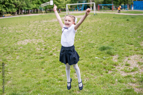 Little blonde schoolgirl in a school uniform with white bows on pigtails, shows a cool sign with her hands, jumps with happiness on a green lawn, she is glad to go to school, laughs toothless smile