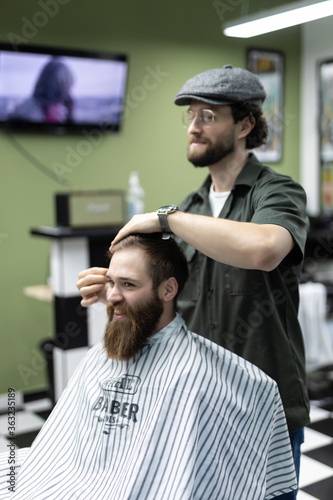 Time for new haircut. Handsome young bearded man looking at his reflection in the mirror and keeping hand in hair while sitting in chair at barbershop