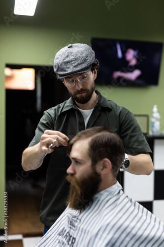 Portrait of attractive young man getting trendy haircut. Male hairdresser serving client, making haircut using metal scissors and comb
