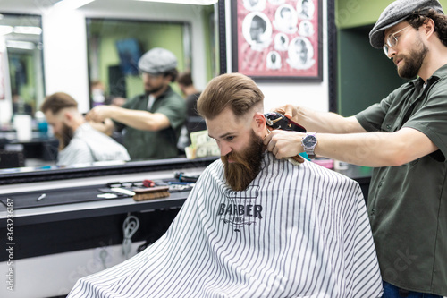 Young barber making haircut with razor to attractive bearded man in barbershop