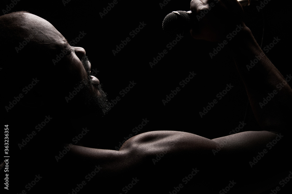 portrait of a dark-skinned muscular handsome guy with a beard on a black background who emotionally sings into a microphone