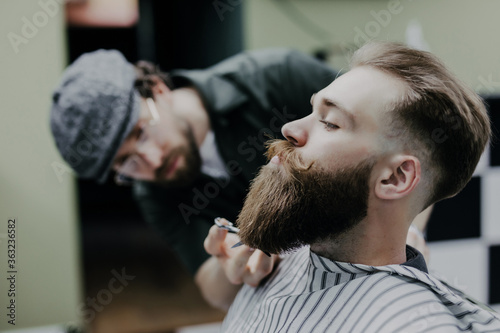 Side view of handsome bearded man having his beard cut by hairdresser at the barbershop