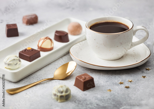 Luxury Chocolate candies in white porcelain plate with cup of black coffee and golden spoon on light table background.