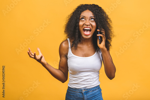 Angry mixed race black african american woman shouting in mobile phone, isolated over yellow background.