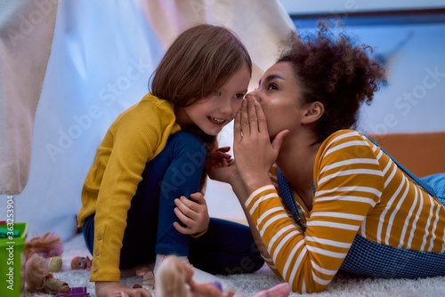 African american woman baby sitter entertaining caucasian cute little girl. They are gossiping and telling secrets sitting in kids room