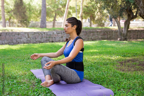 A beautiful girl in a park dressed in sportswear wearing Back posture corrector sit on yoga mat in ypoga pose. Lifestyle, outdoor.