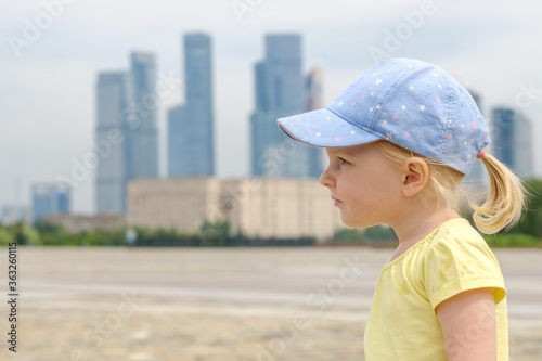 Profile portrait of a blonde little girl baby on a background of skyscrapers. Little kid in a huge city. The girl is wearing a yellow T-shirt and a blue cap. The girl carefully looks into the distance © Nekrasov