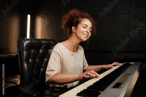 Young pretty woman sensually playing on electric piano and singing a song in recording studio. Attractive musician working in studio