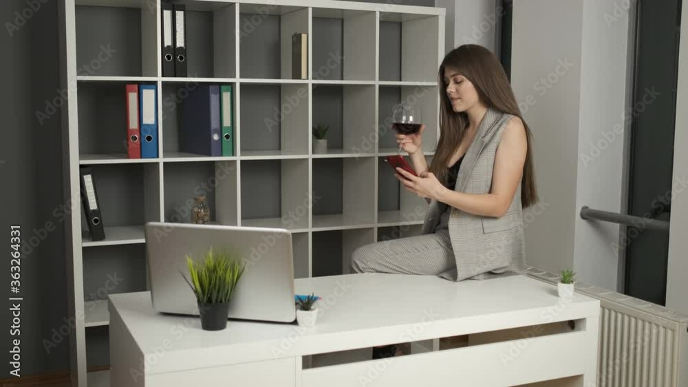 Young beautiful company director sitting in the office on the edge of the table holding a phone in his hands and drinking a glass of red wine after a hard working productive day Young beautiful company director sitting in the office on the edge of the table holding a phone in his hands and drinking a glass of red wine after a hard working productive day