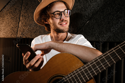 Portrait of attractive male musician with guitar and phone dreamily working in recording studio photo