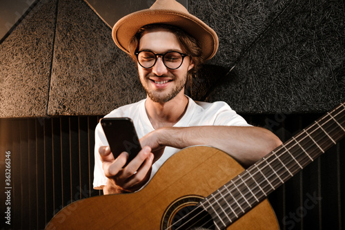 Young attractive man with guitar happily using smartphone in modern sound recording studio photo