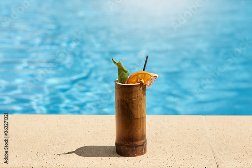 Canvas Print Exotic cocktail in wooden glass with lemon and drinking tube standing on poolside with blue pool's water on background, tasty fresh beverage on resort