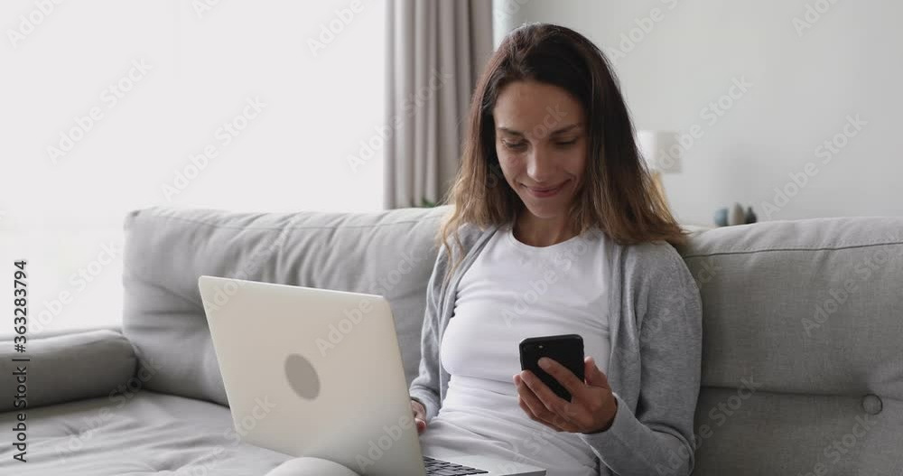 Happy young woman using laptop computer looking at smart cell phone screen laughing enjoying mobile technology internet apps study work with wireless electronic devices online sitting on sofa at home Happy young woman using laptop computer looking at smart cell phone screen laughing enjoying mobile technology internet apps study work with wireless electronic devices online sitting on sofa at home