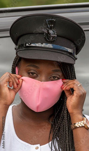 England, UK. 2020.   Portrait of a woman chauffeur with uniform cap and braided hair adjusting her pink coloured facemask during the Covid-19 outbreak photo