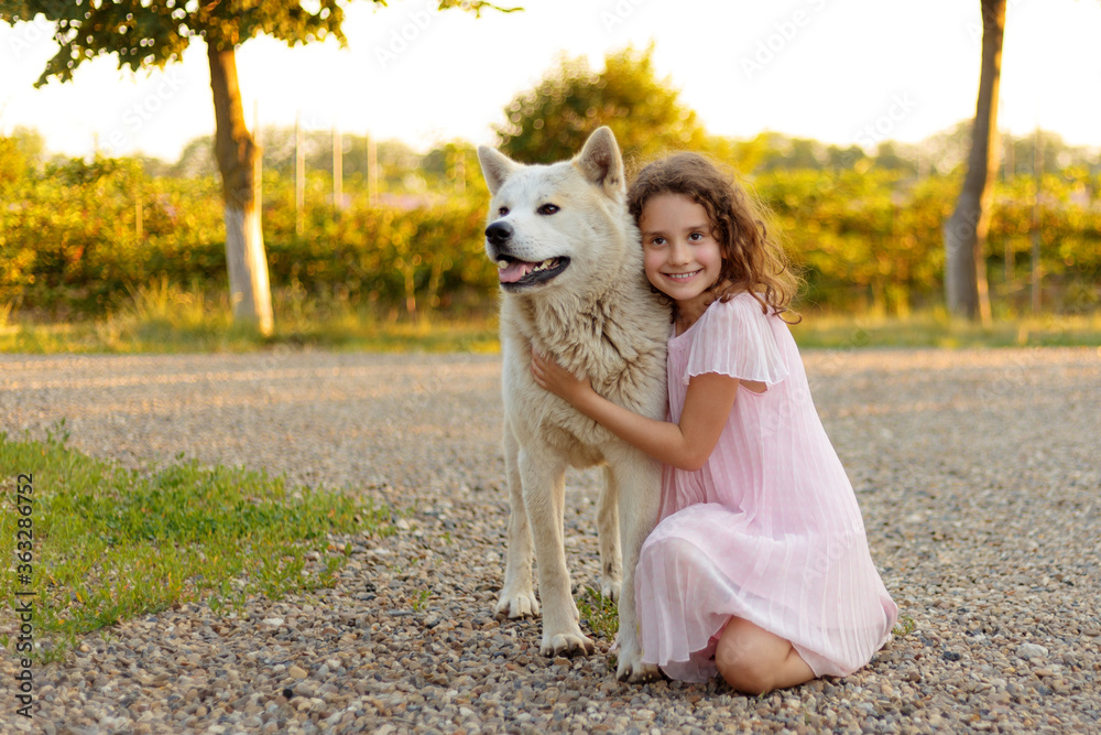Frontal portrait of a little cute girl with a big white dog in the park. A beautiful 7 year old girl in pink dress hugs her favorite dog during a summer walk. Frontal portrait of a little cute girl with a big white dog in the park. A beautiful 7 year old girl in pink dress hugs her favorite dog during a summer walk.