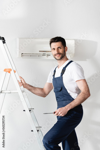 smiling repairman holding screwdriver while standing on stepladder near air conditioner © LIGHTFIELD STUDIOS