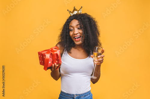 Portrait of an excited young african american black woman holding present box and glass of champagne isolated over yellow background.