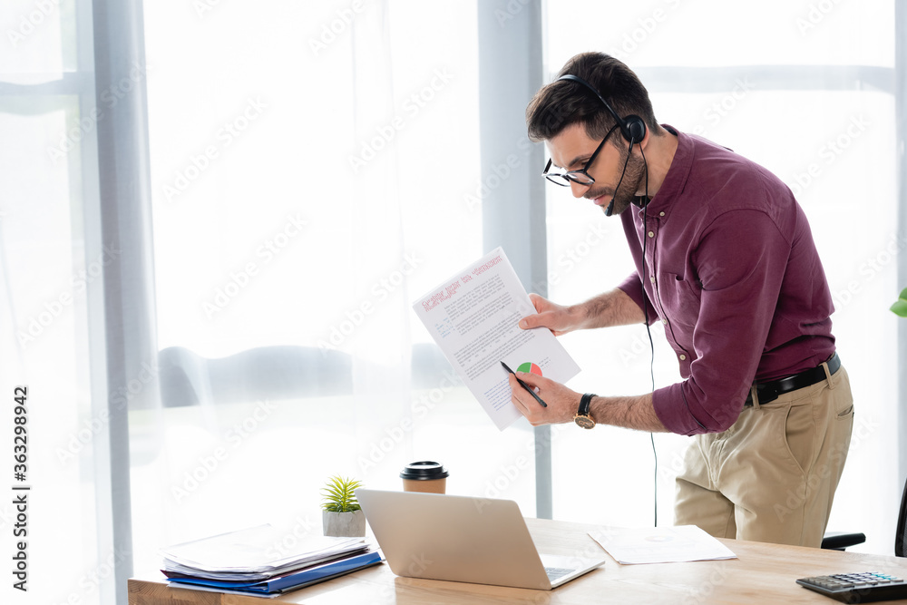 young businessman in headset pointing with pencil at document during online meeting on laptop