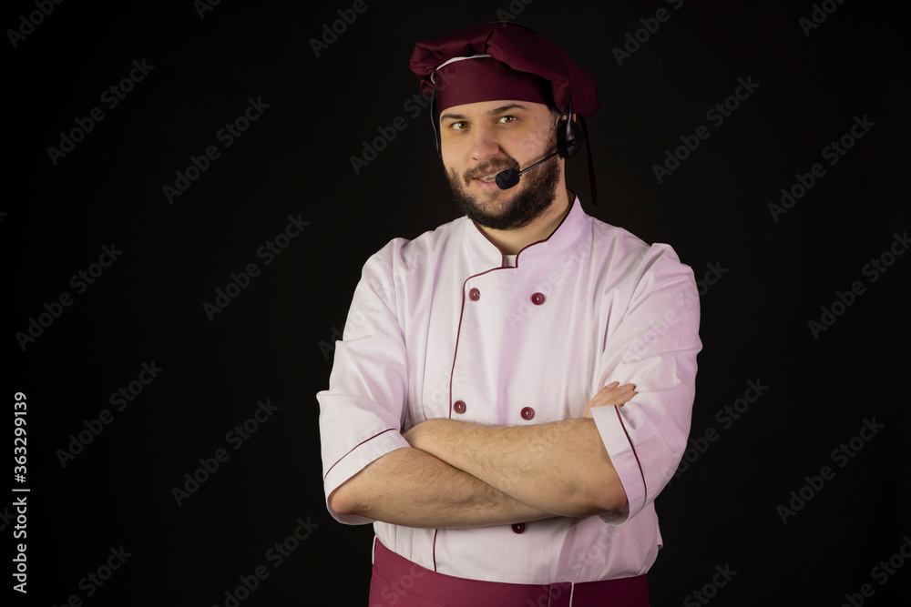 cheerful smiling young bearded male chef cook in uniform wearing headset talking into microphone looking at camera isolated on black. Culinary training concept. Distance management concept. copy space cheerful smiling young bearded male chef cook in uniform wearing headset talking into microphone looking at camera isolated on black. Culinary training concept. Distance management concept. copy space
