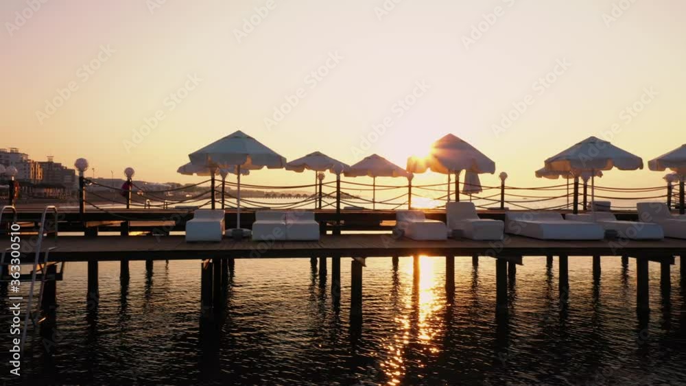 Background of pier at sunset with calm sea water. Marine pier with empty sunbeds and umbrellas in the evening. Seashore vacation concept. Background of pier at sunset with calm sea water. Marine pier with empty sunbeds and umbrellas in the evening. Seashore vacation concept.